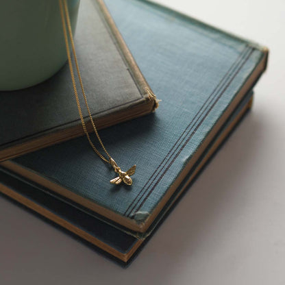 18ct gold bee necklace on fine chain, flatlay jewellery photo, on stacked books 